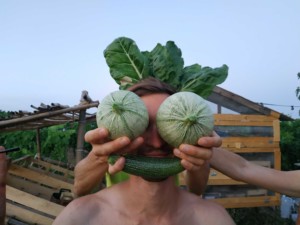 Une personne devant le visage de laquelle sont tenus deux courgettes rondes en guise d'yeux, une courgette droite en guise de bouche et des feuilles de chou en guis de cheveux